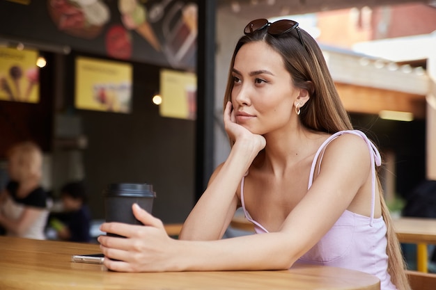 Giovane ragazza graziosa asiatica alla moda attraente che si siede alla tavola calda con la tazza di caffè