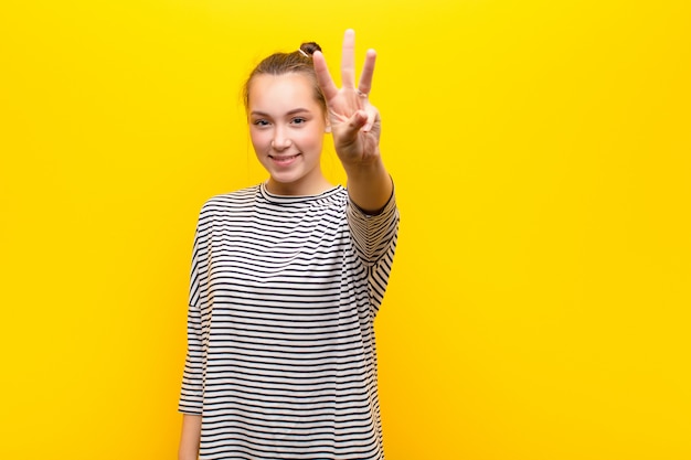 Giovane ragazza graziosa bionda che sorride e che sembra amichevole, mostrando il numero due o il secondo con la mano in avanti, contando contro il muro giallo