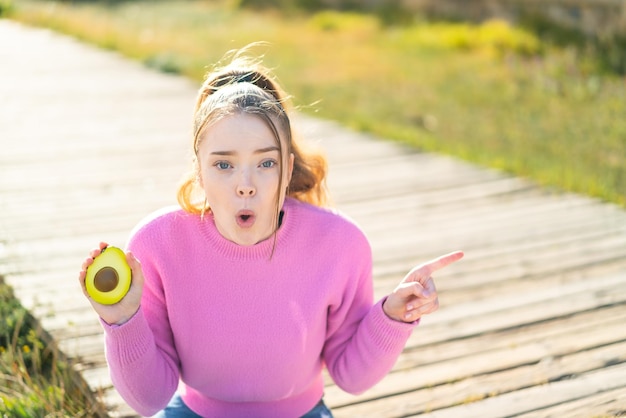 Giovane ragazza graziosa che tiene un avocado all'aperto sorpreso e che indica il lato