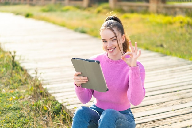 Giovane ragazza graziosa che tiene una compressa all'aperto che mostra il segno giusto con le dita