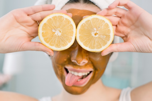 Giovane ragazza graziosa con la maschera di fango marrone sul viso che tiene le metà della frutta del limone, coprendo gli occhi. Ragazza teenager che si prende cura della sua pelle. Trattamento di bellezza.