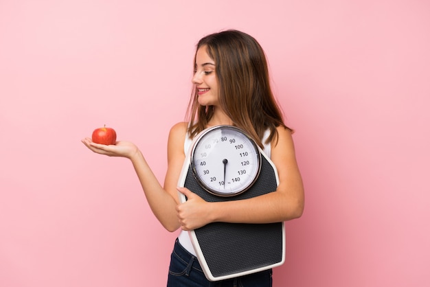 Giovane ragazza graziosa con la pesa e con una mela sopra la parete rosa isolata