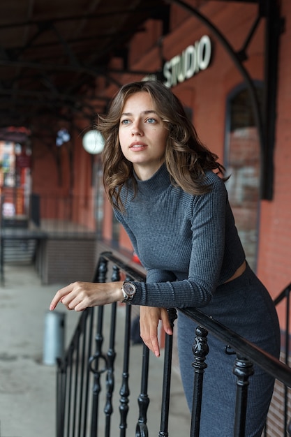 Giovane ragazza graziosa premurosa in dolcevita grigio, gonna con orologi sul braccio pone davanti all'edificio rosso