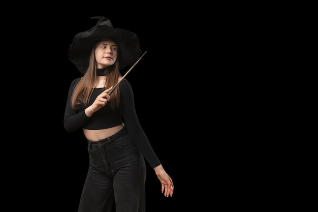 Giovane ragazza in abiti neri e cappello appuntito utilizza bacchetta magica sfondo nero Costume da strega di Halloween