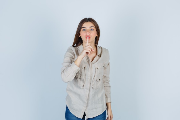 Giovane ragazza in camicia beige, jeans che mostrano gesto di silenzio e sembrano seri, vista frontale.