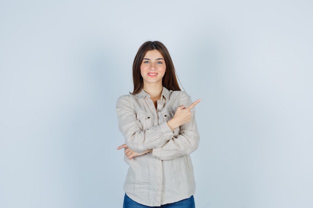 Giovane ragazza in camicia beige, jeans che punta a destra con il dito indice e sembra felice, vista frontale.