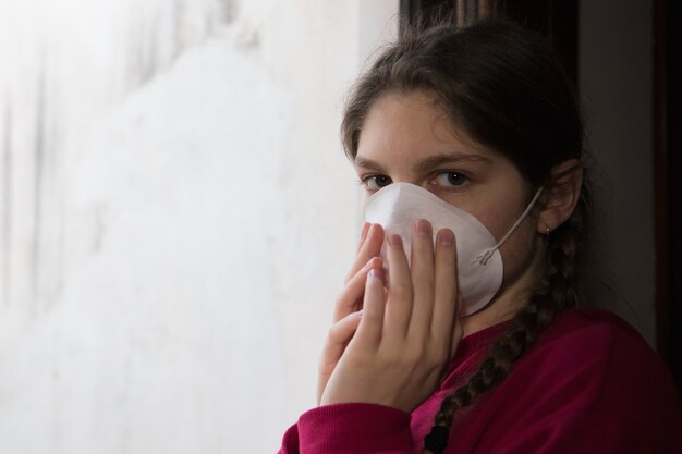 Giovane ragazza in isolamento domestico, durante la quarantena indossa una maschera protettiva per evitare la predicazione della malattia coronavirus, vicino alla finestra