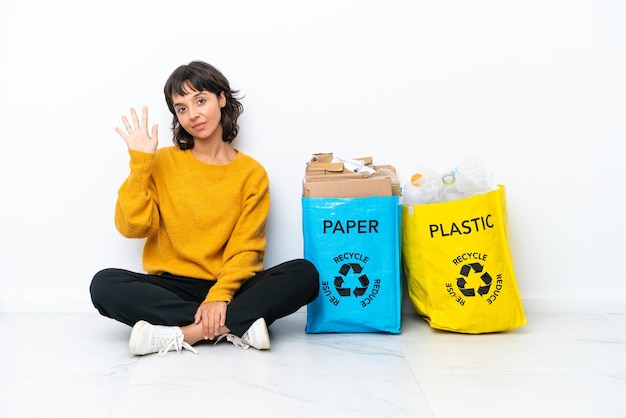 Giovane ragazza in possesso di un sacchetto pieno di plastica e carta seduta sul pavimento isolato su sfondo bianco contando cinque con le dita