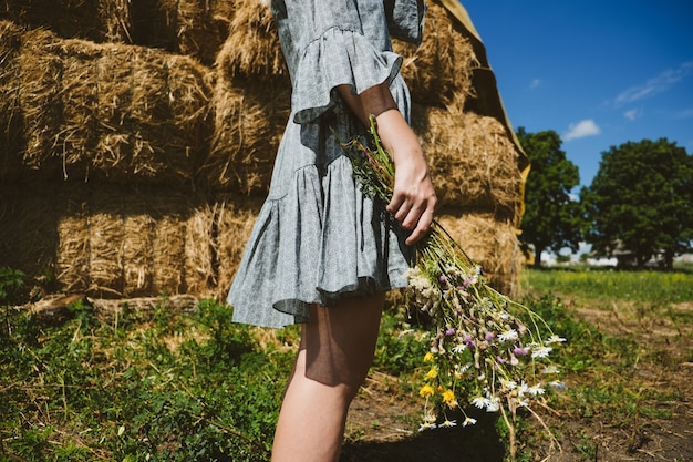 Giovane ragazza in stivali di gomma con fiori in piedi sullo sfondo di balle di paglia in campagna