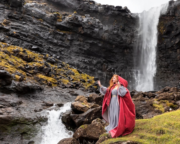 Giovane ragazza in un mantello rosso rimane vicino alla cascata di Fossa in autunno, Streymoy, Isole Faerøer