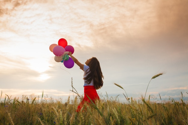 Giovane ragazza in una gonna rossa in esecuzione con palloncini colorati nel campo di grano. Ispirazione alla natura, retroilluminazione
