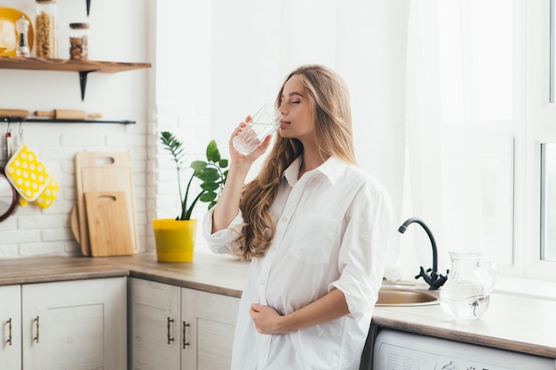 Giovane ragazza incinta carina che beve acqua pulita in cucina