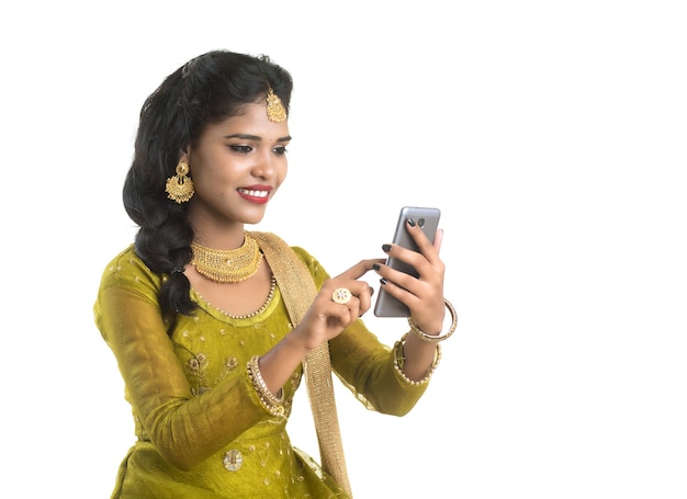 Giovane ragazza indiana tradizionale utilizzando un telefono cellulare o uno smartphone isolato su un muro bianco