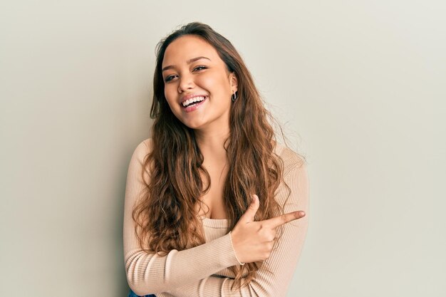 Giovane ragazza ispanica che indossa abiti casual sorridendo allegro indicando con la mano e il dito fino al lato