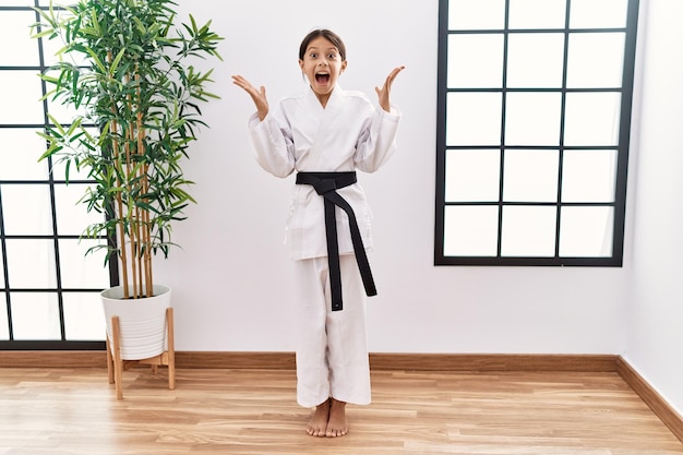 Giovane ragazza ispanica che indossa kimono da karate e cintura nera che festeggia pazza e stupita per il successo con le braccia alzate e gli occhi aperti che urlano eccitati. concetto di vincitore