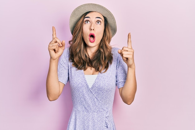 Giovane ragazza ispanica che indossa un cappello estivo stupito e sorpreso guardando in alto e indicando con le dita e le braccia alzate.