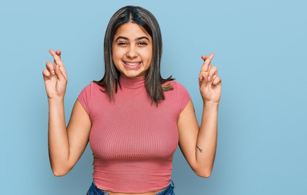 Giovane ragazza ispanica che indossa una maglietta casual che gesturing dito incrociato sorridendo con speranza e occhi chiusi fortuna e concetto superstizioso