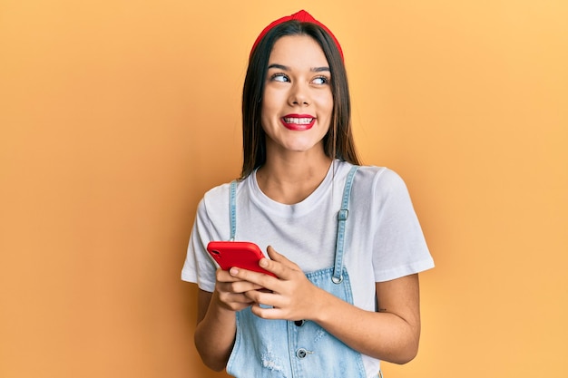 Giovane ragazza ispanica che usa lo smartphone sorridendo guardando di lato e fissando via il pensiero