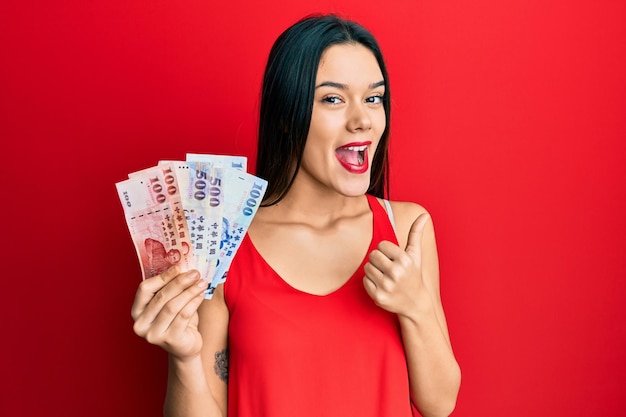 Giovane ragazza ispanica con nuove banconote in dollari taiwanesi che indica il pollice verso il lato sorridendo felice con la bocca aperta