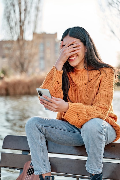 Giovane ragazza latina che guarda il suo smartphone con la mano sul viso coprendosi gli occhi con imbarazzo sorpresa e una faccia felice concetto di tecnologia e relazioni sociali