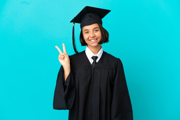 Giovane ragazza laureata sopra fondo blu isolato che sorride e che mostra il segno di vittoria