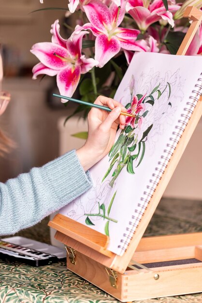 Giovane ragazza mancina che dipinge natura morta con fiori gigli viola con colori ad acquerello sul cavalletto