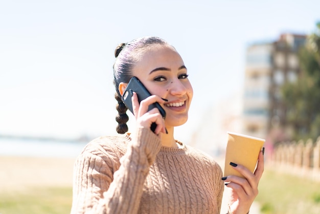 Giovane ragazza marocchina all'aperto utilizzando il telefono cellulare e tenendo un caffè con un'espressione felice