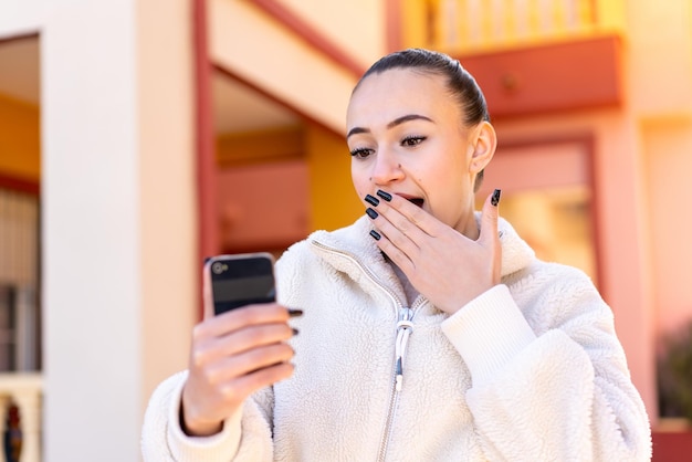 Giovane ragazza marocchina che utilizza il telefono cellulare all'aperto con espressione facciale sorpresa e scioccata