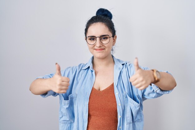 Giovane ragazza moderna con i capelli blu in piedi sopra il segno di successo su sfondo bianco che fa un gesto positivo con la mano pollice in alto sorridente e felice espressione allegra e gesto del vincitore