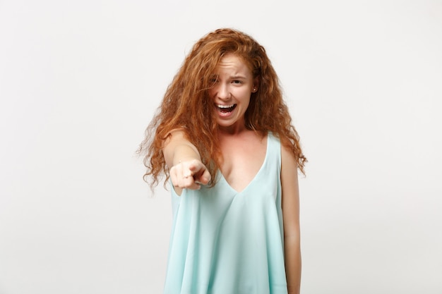 Giovane ragazza rossa irritata della donna in vestiti leggeri casuali che posano isolati sul ritratto bianco dello studio del fondo. Concetto di stile di vita della gente. Mock up copia spazio. Puntare il dito indice sulla fotocamera, imprecando.