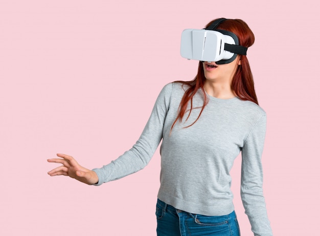 Giovane ragazza rossa utilizzando occhiali VR. Esperienza di realtà virtuale su sfondo rosa isolato