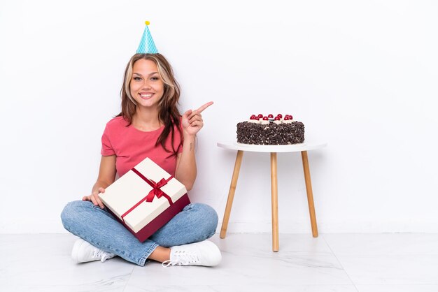 Giovane ragazza russa che celebra un compleanno seduto sul pavimento isolato su sfondo bianco che punta il dito di lato