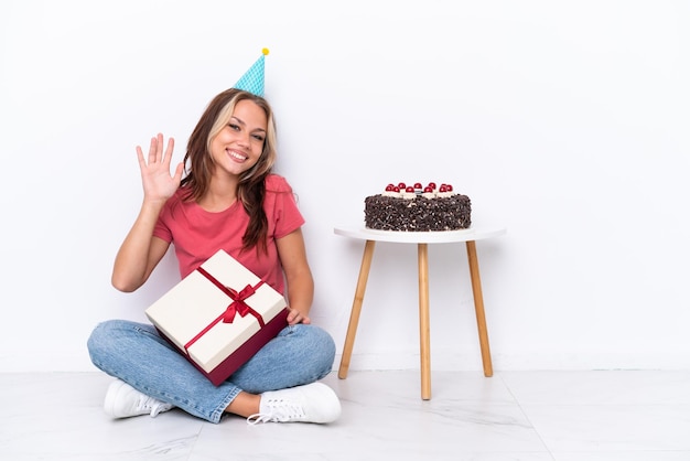 Giovane ragazza russa che celebra un compleanno seduto sul pavimento isolato su sfondo bianco salutando con la mano con un'espressione felice