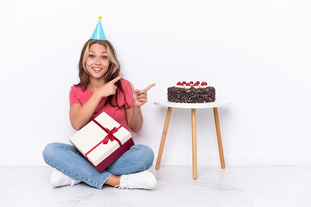 Giovane ragazza russa che festeggia un compleanno seduta sul pavimento isolato su sfondo bianco sorpresa e che indica il lato