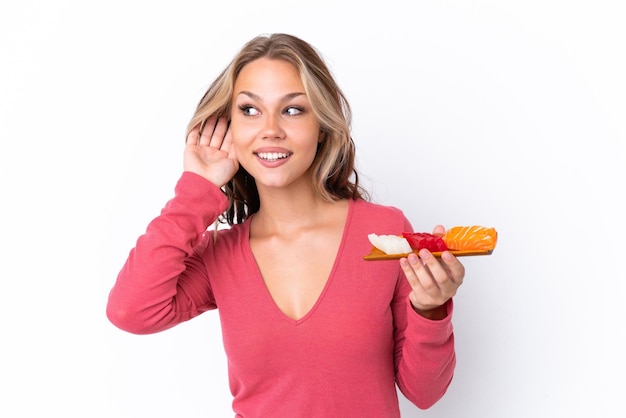 Giovane ragazza russa che tiene sashimi isolato su sfondo bianco ascoltando qualcosa mettendo la mano sull'orecchio