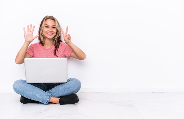 Giovane ragazza russa con un computer portatile seduto sul pavimento isolato su sfondo bianco contando sette con le dita