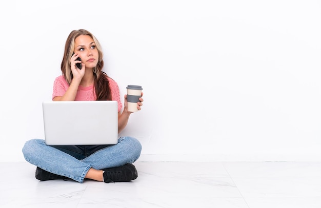 Giovane ragazza russa con un computer portatile seduto sul pavimento isolato su sfondo bianco tenendo il caffè da portare via e un cellulare