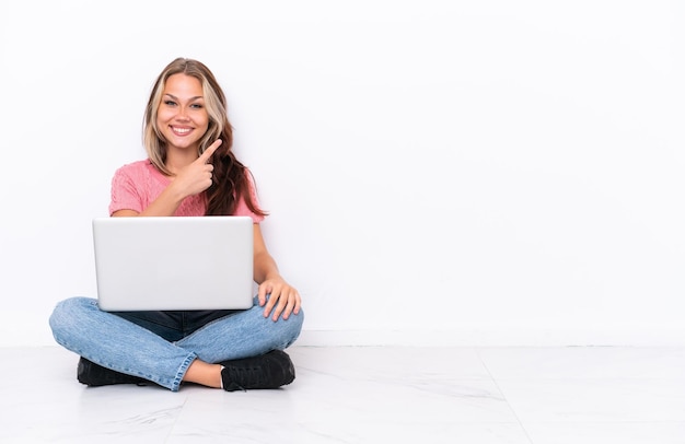 Giovane ragazza russa con un laptop seduto sul pavimento isolato su sfondo bianco che indica il lato per presentare un prodotto