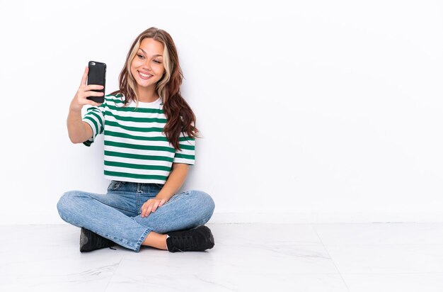 Giovane ragazza russa seduta sul pavimento isolata su sfondo bianco che fa un selfie