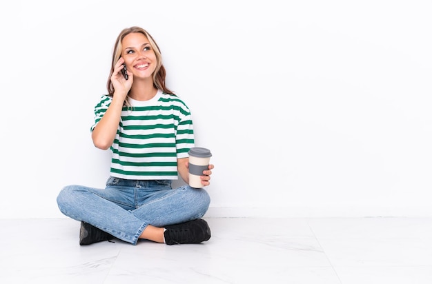 Giovane ragazza russa seduta sul pavimento isolata su sfondo bianco con in mano un caffè da asporto e un cellulare