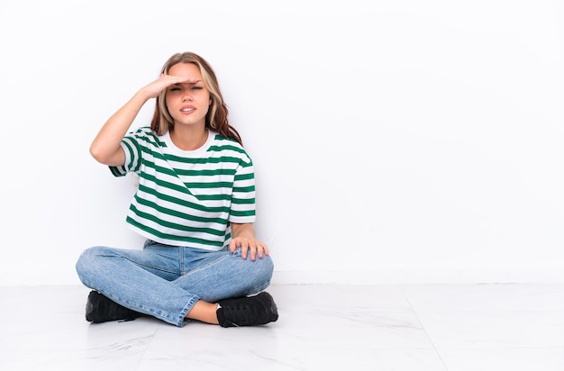 Giovane ragazza russa seduta sul pavimento isolata su sfondo bianco guardando lontano con la mano per guardare qualcosa