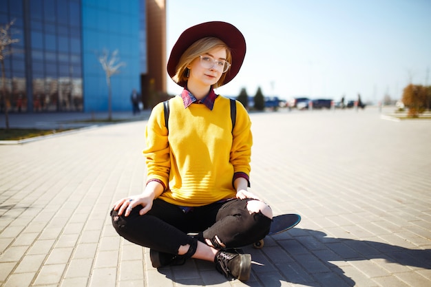 Giovane ragazza seduta su uno skateboard e mandare SMS su un telefono cellulare. Ragazza hipster in un maglione giallo,