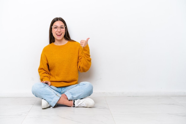 Giovane ragazza seduta sul pavimento isolata su sfondo bianco che punta di lato per presentare un prodotto