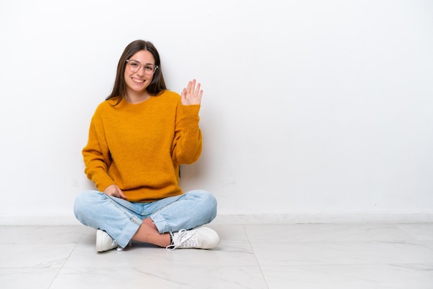 Giovane ragazza seduta sul pavimento isolato su sfondo bianco salutando con la mano con l'espressione felice