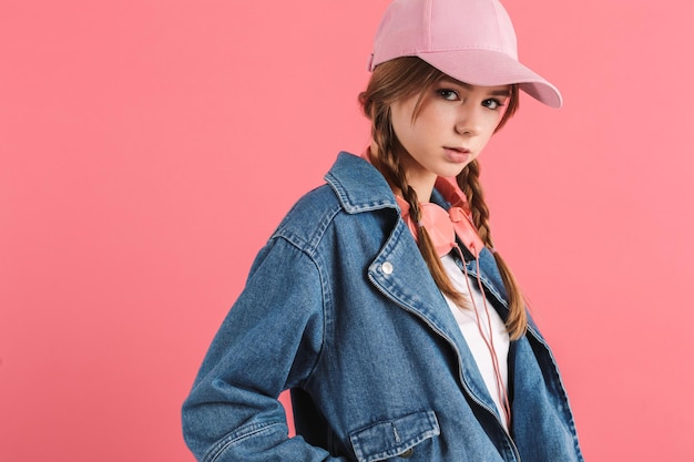 Giovane ragazza seria con due trecce in giacca di jeans e berretto con cuffie sul collo che guarda premurosamente a porte chiuse su sfondo rosa isolato