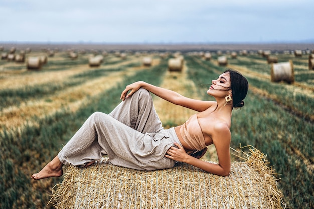 Giovane ragazza snella del brunette, trovandosi sul fieno, proponendo per la macchina fotografica, tiro della foto sul campo, cielo blu.