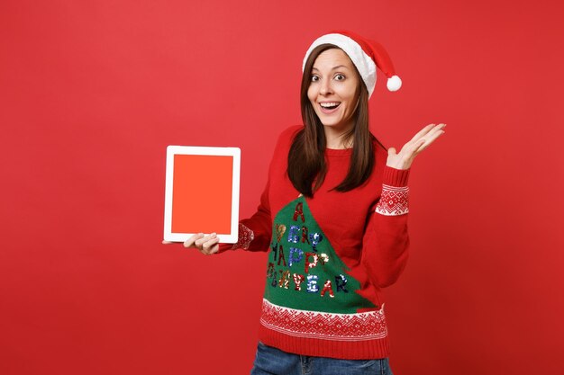 Giovane ragazza sorpresa di Santa che spande le mani, tenendo il computer tablet pc con schermo vuoto vuoto isolato su sfondo rosso. Felice Anno Nuovo 2019 celebrazione festa concetto. Mock up copia spazio.