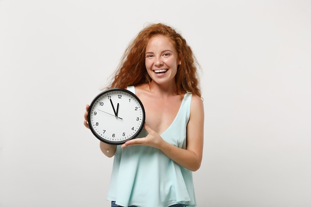 Giovane ragazza sorridente allegra carina rossa della donna in vestiti leggeri casuali che posano isolata sul fondo bianco della parete, ritratto dello studio Concetto di stile di vita della gente. Mock up copia spazio. Tenendo l'orologio rotondo.