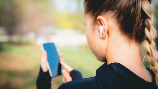 Giovane ragazza sorridente che fa sport e corre nel parco usando il suo telefono per ascoltare la musica con le cuffie wireless al tramonto in città guardando lo schermo