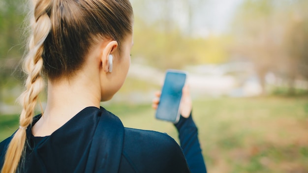 Giovane ragazza sorridente che fa sport e corre nel parco usando il suo telefono